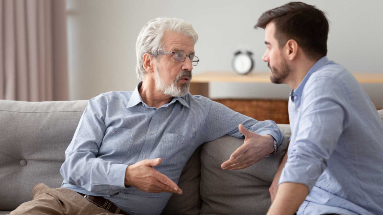 Serious 60s elderly father and grown up adult son sitting on sofa talking having important conversation trying to solve life issues problem, different men relative people communication at home concept