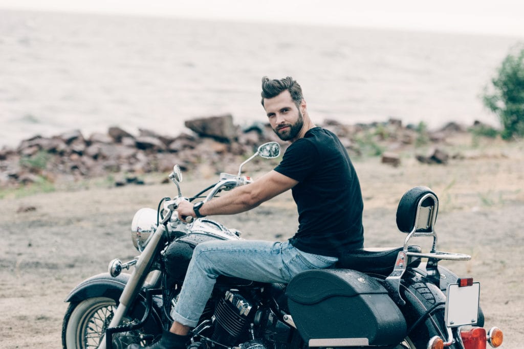 handsome bearded motorcyclist on black motorcycle at sandy beach near sea