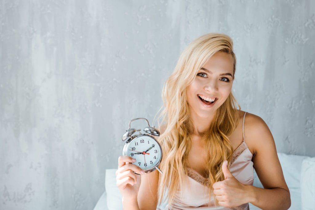 Happy Young Woman Holding Alarm Clock And Smiling At Camera In Bedroom Stockpack Deposit Photos