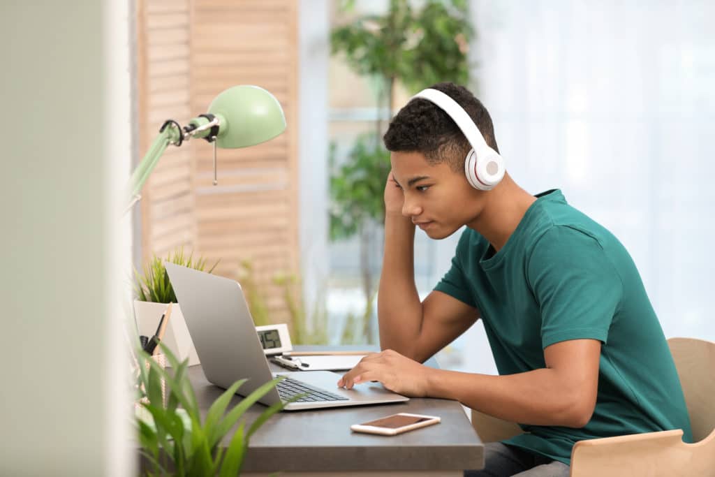 African American Teenage Boy With Headphones Using Laptop At Table In