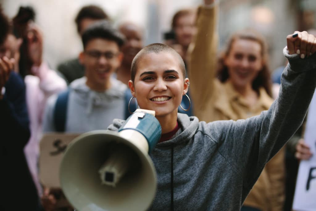 Young Woman Standing Outdoors With Group Of Demonstrator In Background.