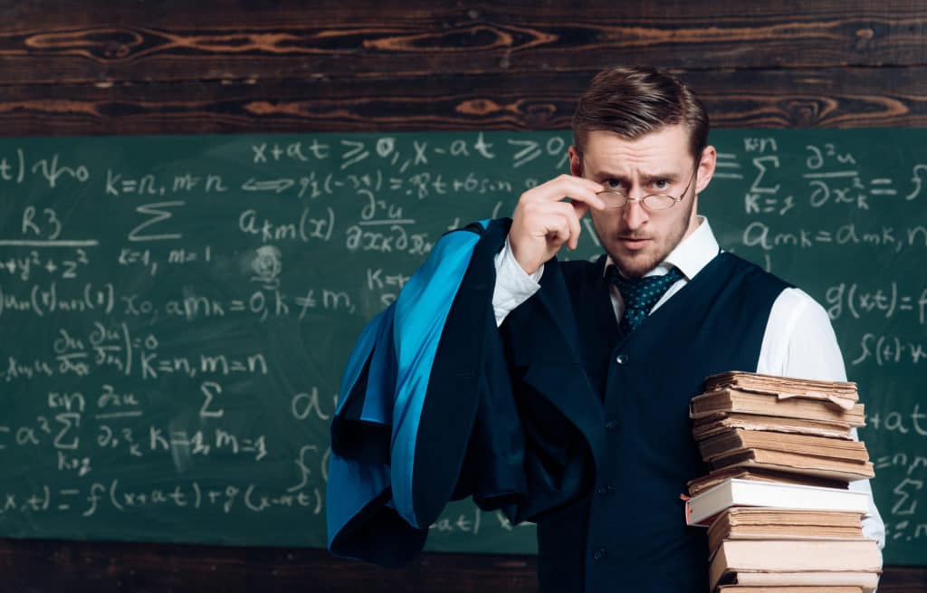 Young Professor Holding Pile Of Books Looking Over His Glasses.