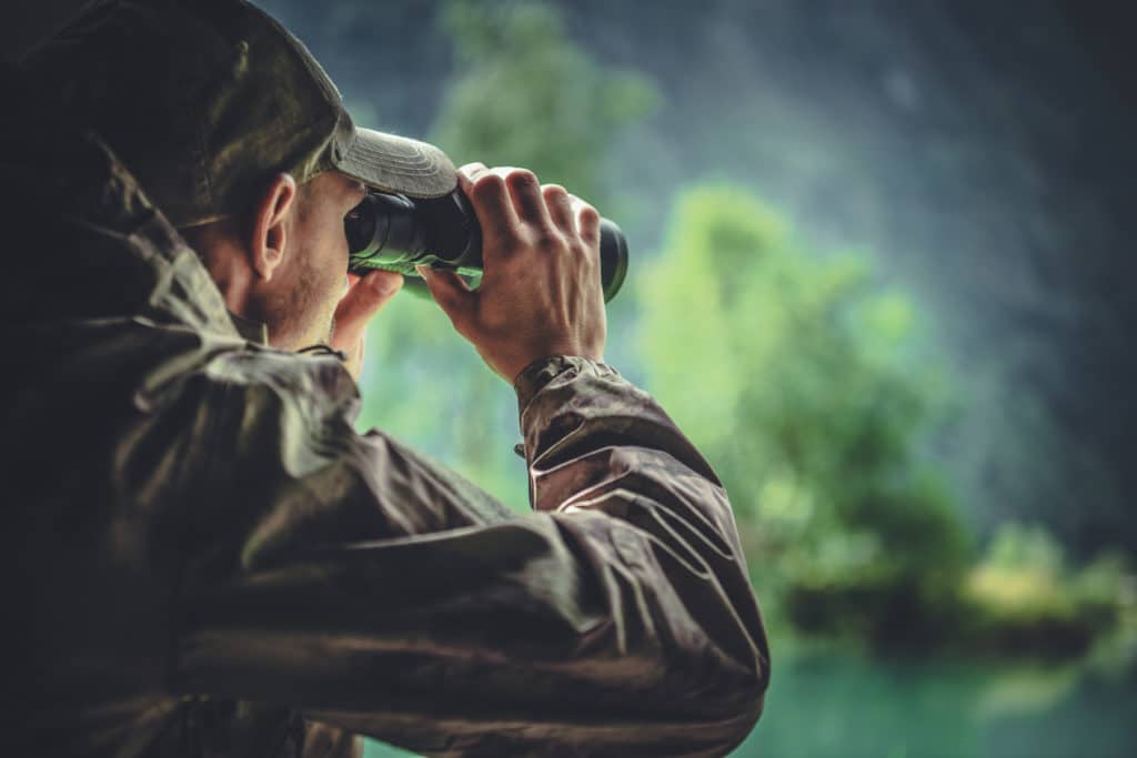 Caucasian Hunter In Masking Camouflage Uniform With Binoculars. Hunter Spotting