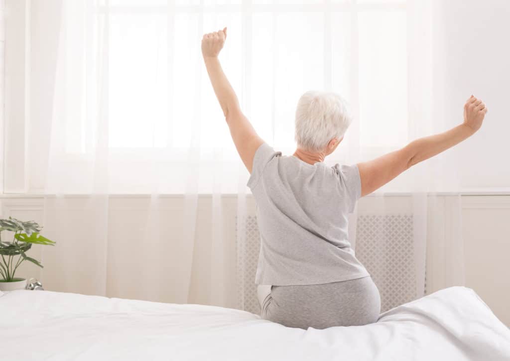 Senior Woman Sitting On Her Bed In Morning Stretching With