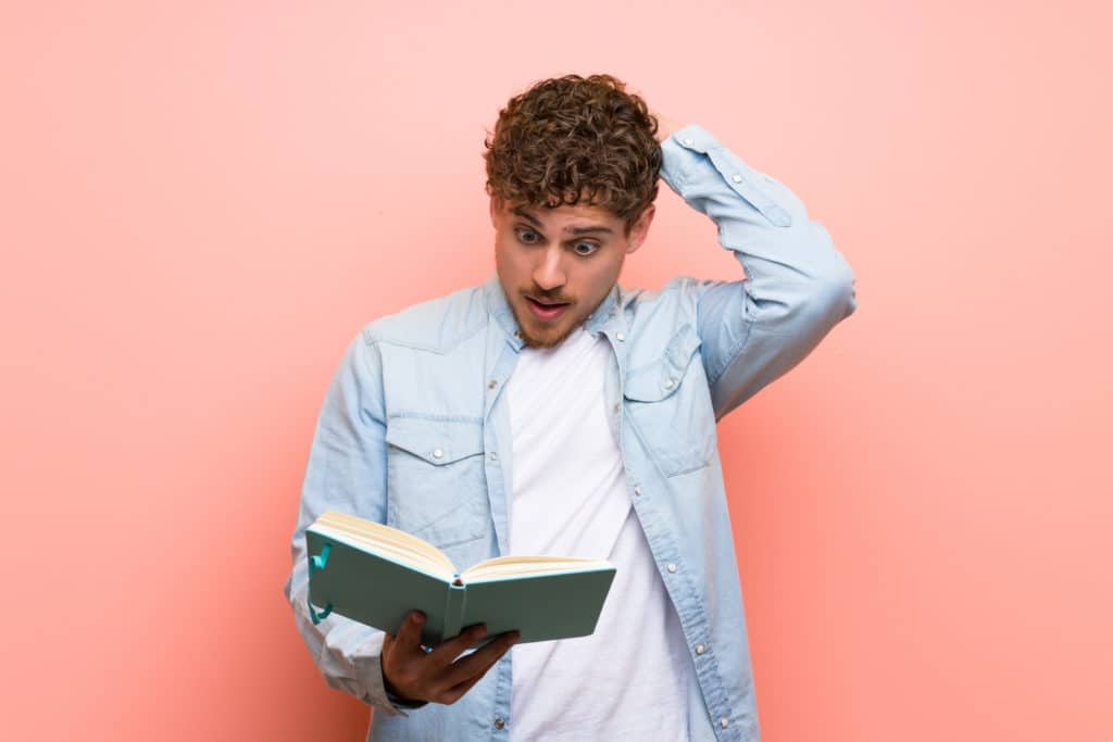 Blonde Man Over Pink Wall Surprised While Enjoying Reading A