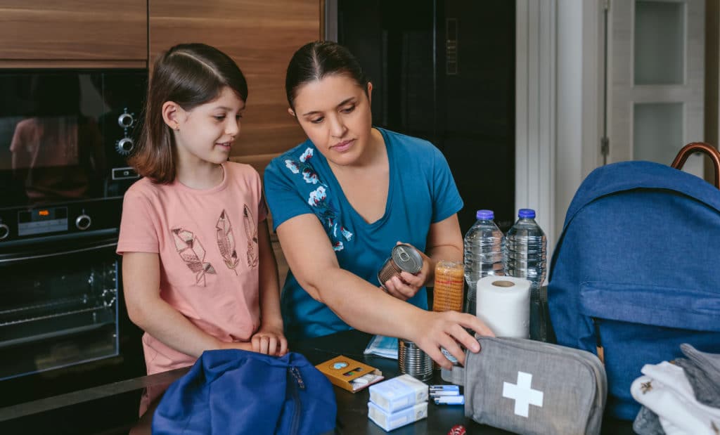Mother Preparing Emergency Backpack With Her Daughter In The Kitchen