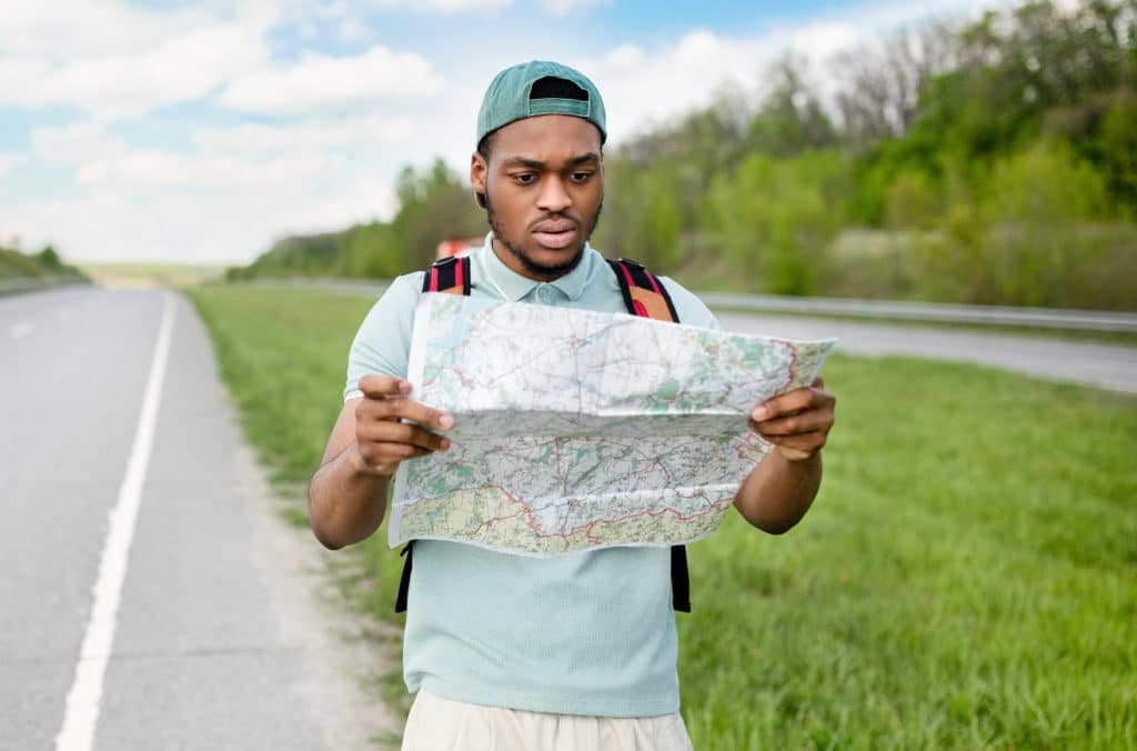 Unhappy Young Black Man Hitchhiking On Road Looking At Map 