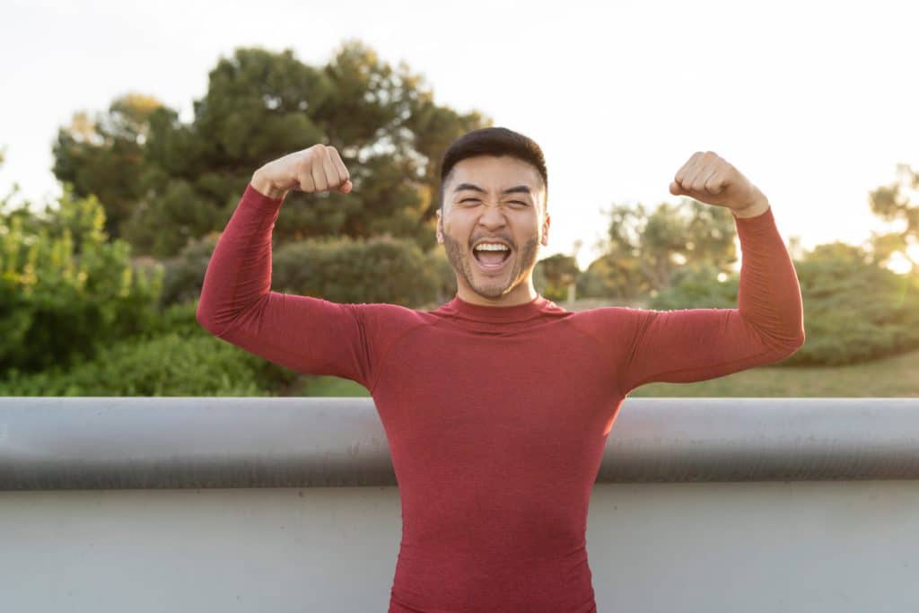 Muscular Asian Man Flexing Biceps With Both Arms Outdoors At
