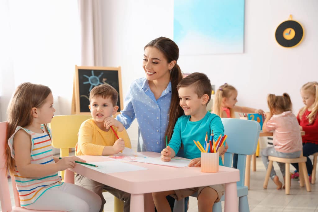 Little Children With Kindergarten Teacher Drawing At Table Indoors. Learning