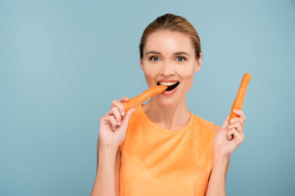 Pleased Woman Looking At Camera While Eating Whole Carrot Isolated