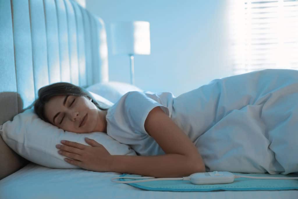 Young Woman Sleeping On Electric Heating Pad In Bed At