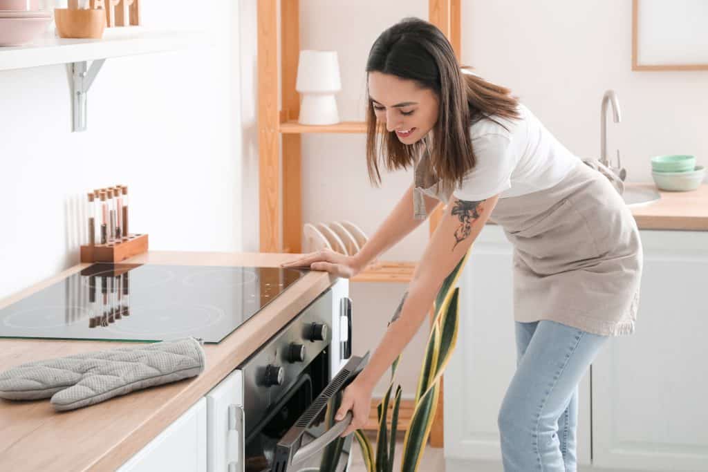 Young Woman Opening Electric Oven In Kitchen